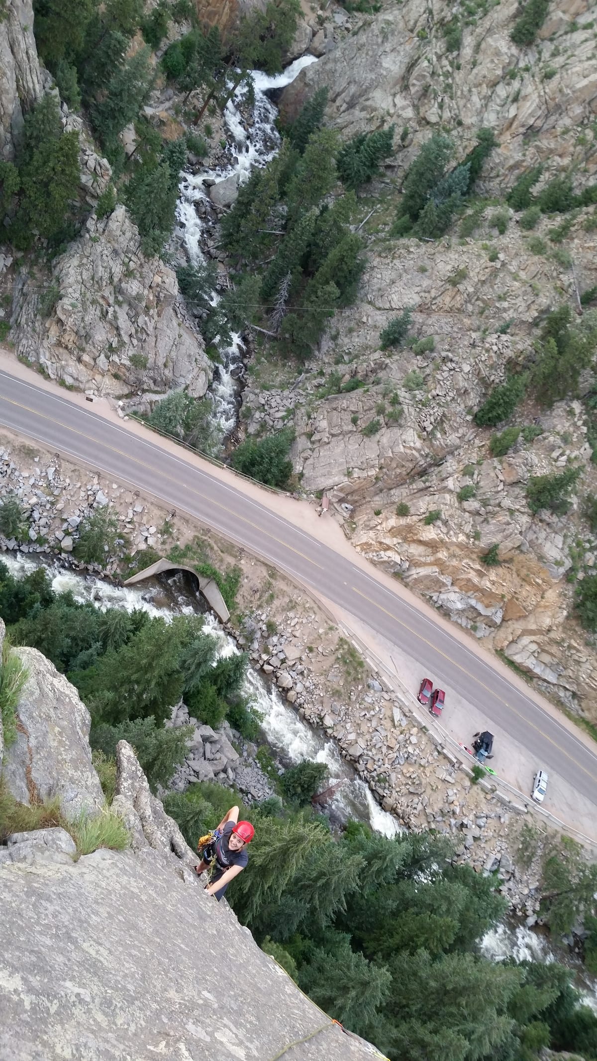 A climber on a steep rock face above a winding canyon road.