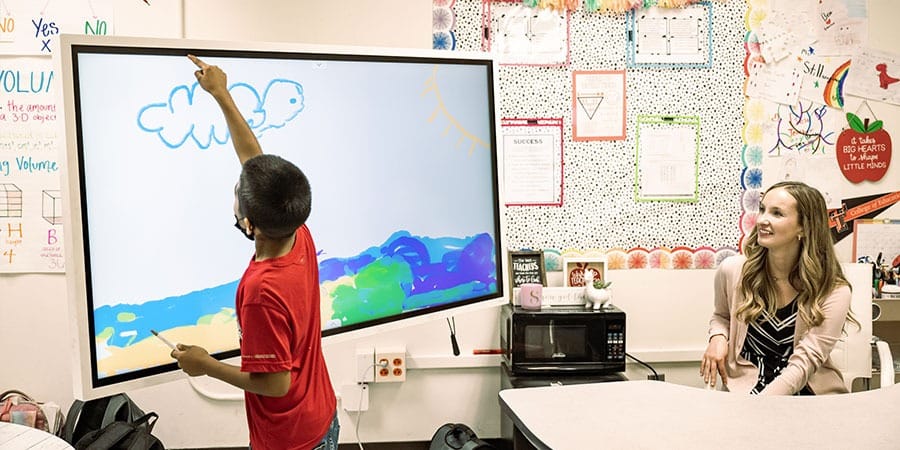 A student draws on a Samsung interactive display beside a teacher in a classroom.